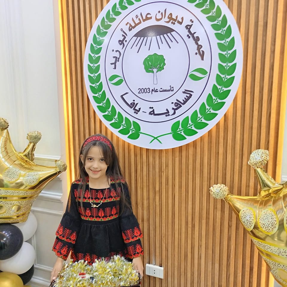 طفلة بالثوب الفلسطيني أمام شعار جمعية ديوان عائلة أبو زيد في حفل الافتتاح